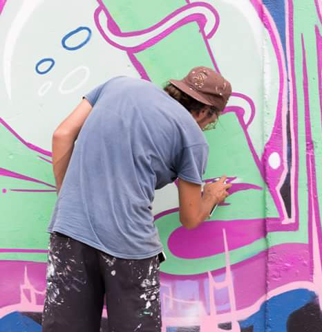 A person is spray-painting colorful graffiti on a wall, predominantly featuring pink and green colors. The individual is seen from the back, wearing a cap and a paint-splattered shirt.