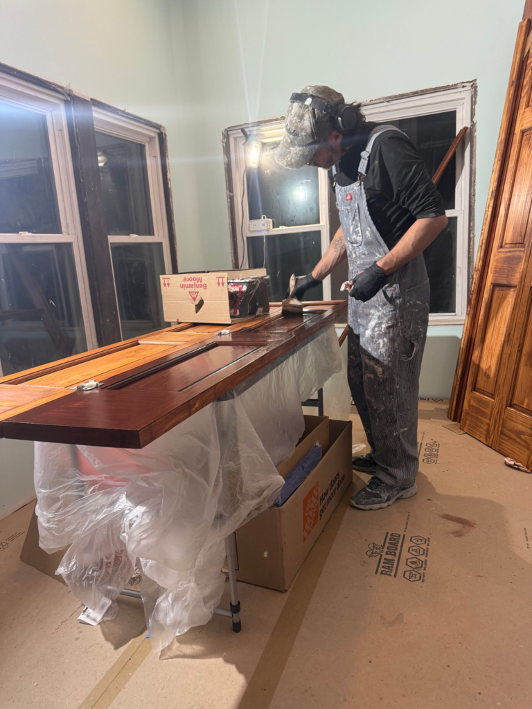 A person is applying finish to a wooden surface in a room with large windows. They are wearing gloves and a cap, and the workspace is covered with protective plastic. Various supplies are visible on the worktable.