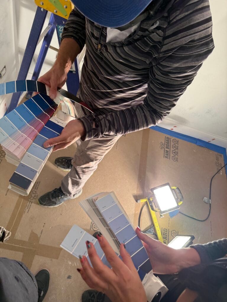 Two people are holding color swatches in a construction setting, with a ladder and work lights in the background.