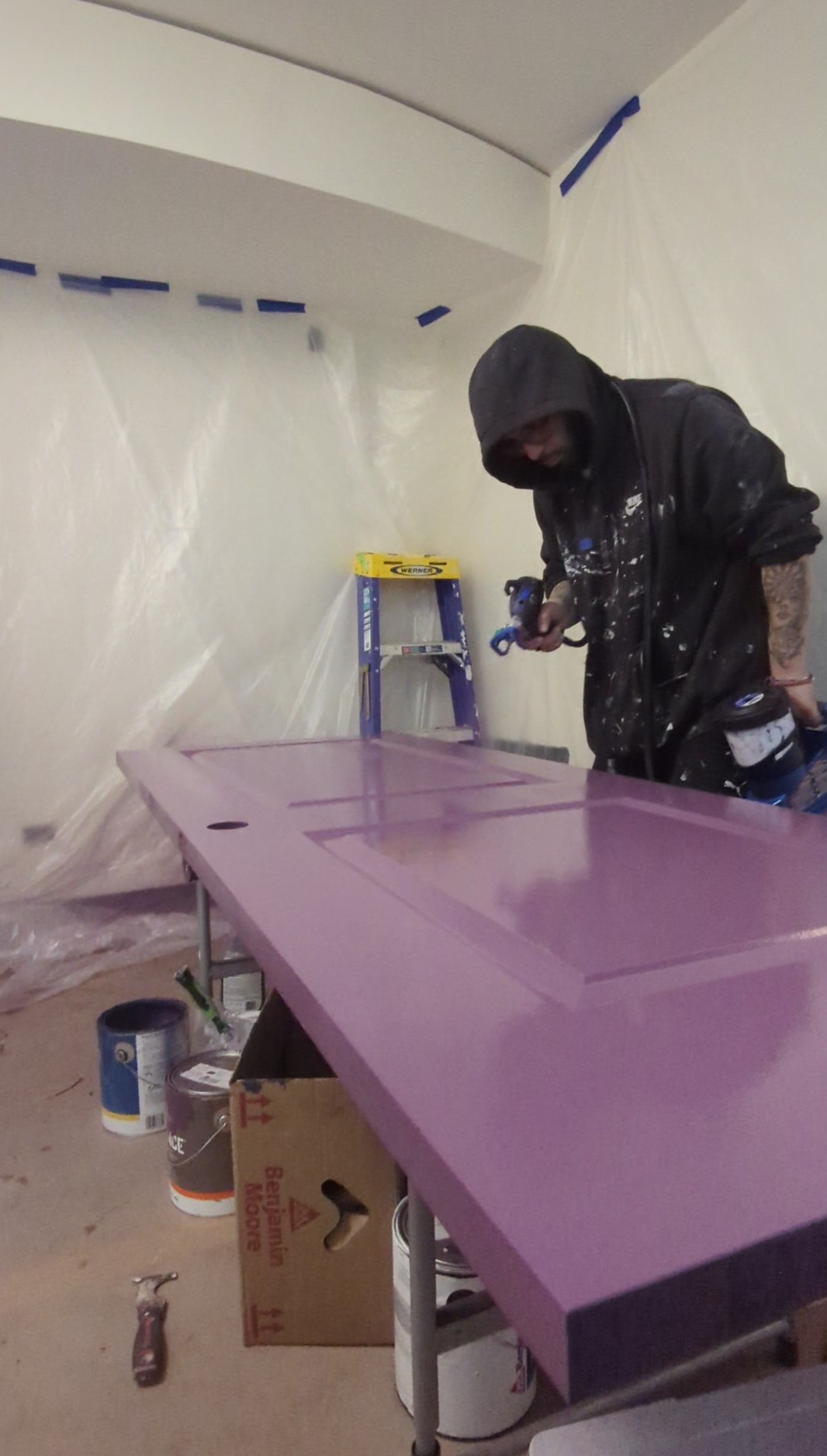 A person wearing a hoodie is spray painting a purple door in a plastic-covered room. Paint cans and a ladder are visible in the background.